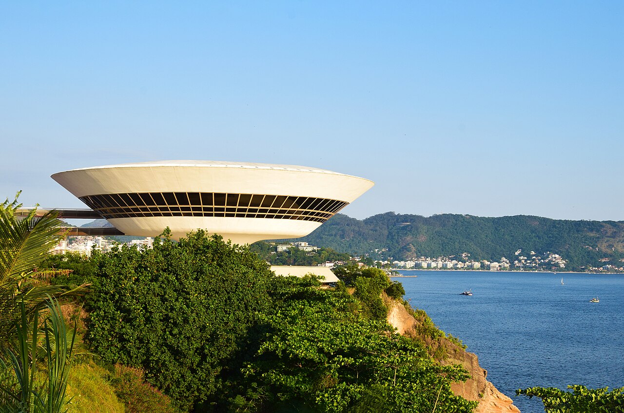 Museu de Arte Contemporânea de Niterói no mirante da Boa Viagem, cercado de vegetação, com a Baía de Guanabara e morros ao fundo